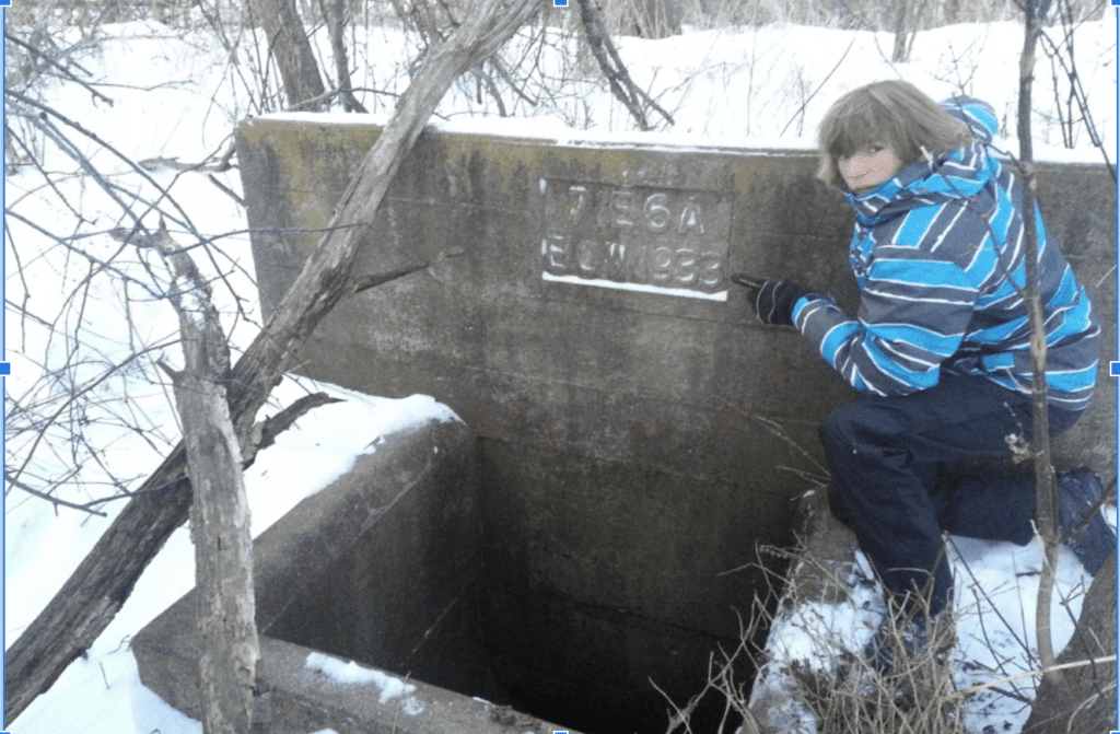 A person in a blue-striped winter jacket kneels next to a concrete structure with an engraved plaque, reminiscent of the rustic charm found at Doudlah Farms. The structure has an open, dark entrance and is surrounded by snow and bare branches, capturing the essence of organic beauty.